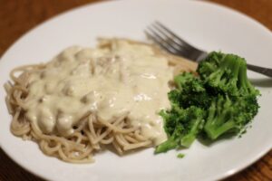 homemade alfredo sauce served over noodles and chicken on a plate. broccoli on side.
