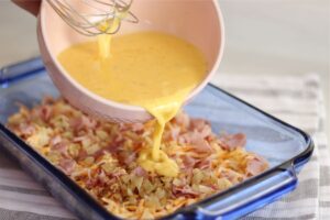 pouring egg mixture from mixing bowl on top of layered ingredients of breakfast casserole