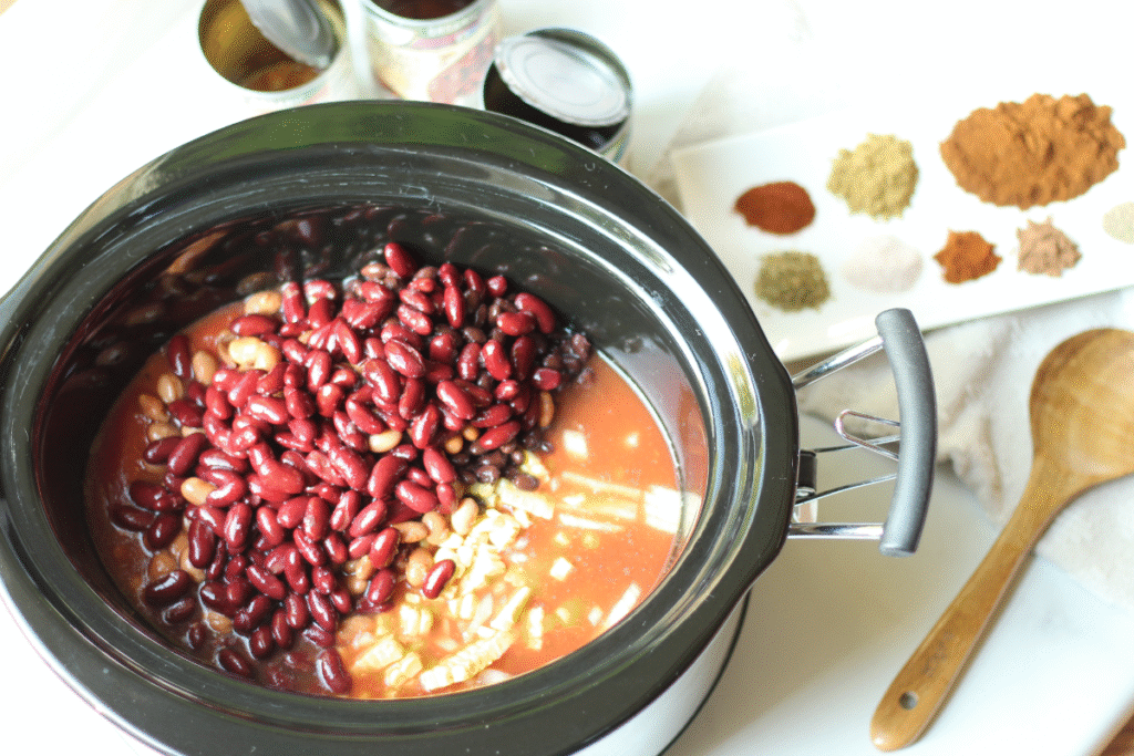 crockpot shown with some ingredients added for best homemade chili recipe. spices and wooden spoon alongside.