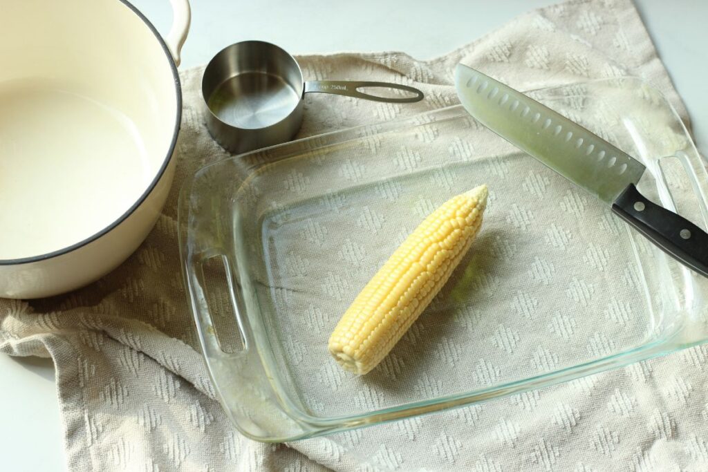 ear of fresh sweet corn in baking dish with knife, measuring cup, and large pot alongside