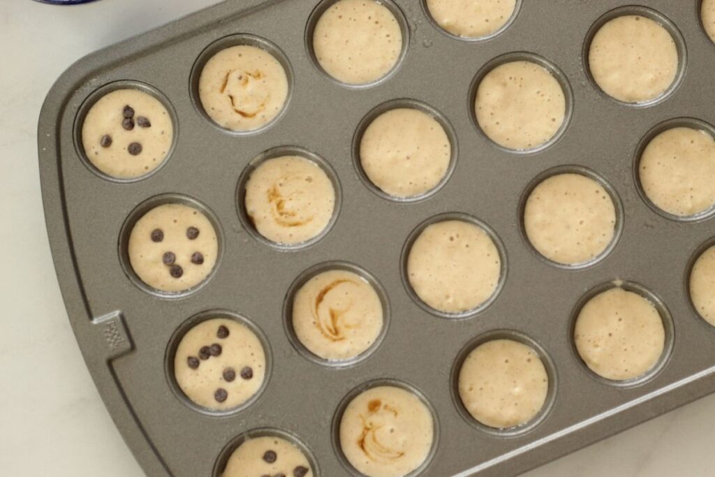 muffin tin filled with mini pancake muffin batter. some cavities have optional add-ins.